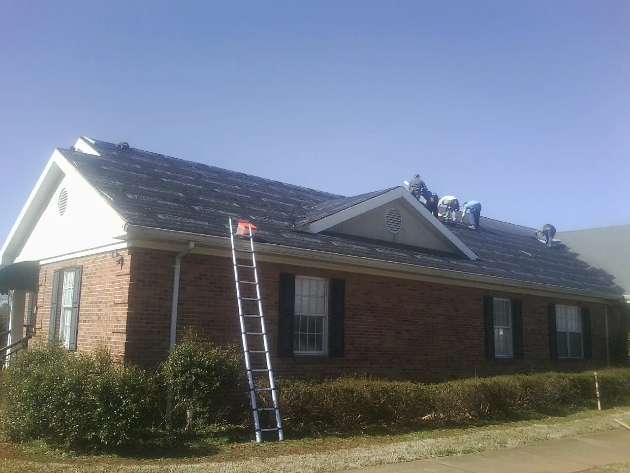 Crew tearing shingles off a brick ranch roof with a ladder set — Metal Roof Repair work by our crew in Lansford